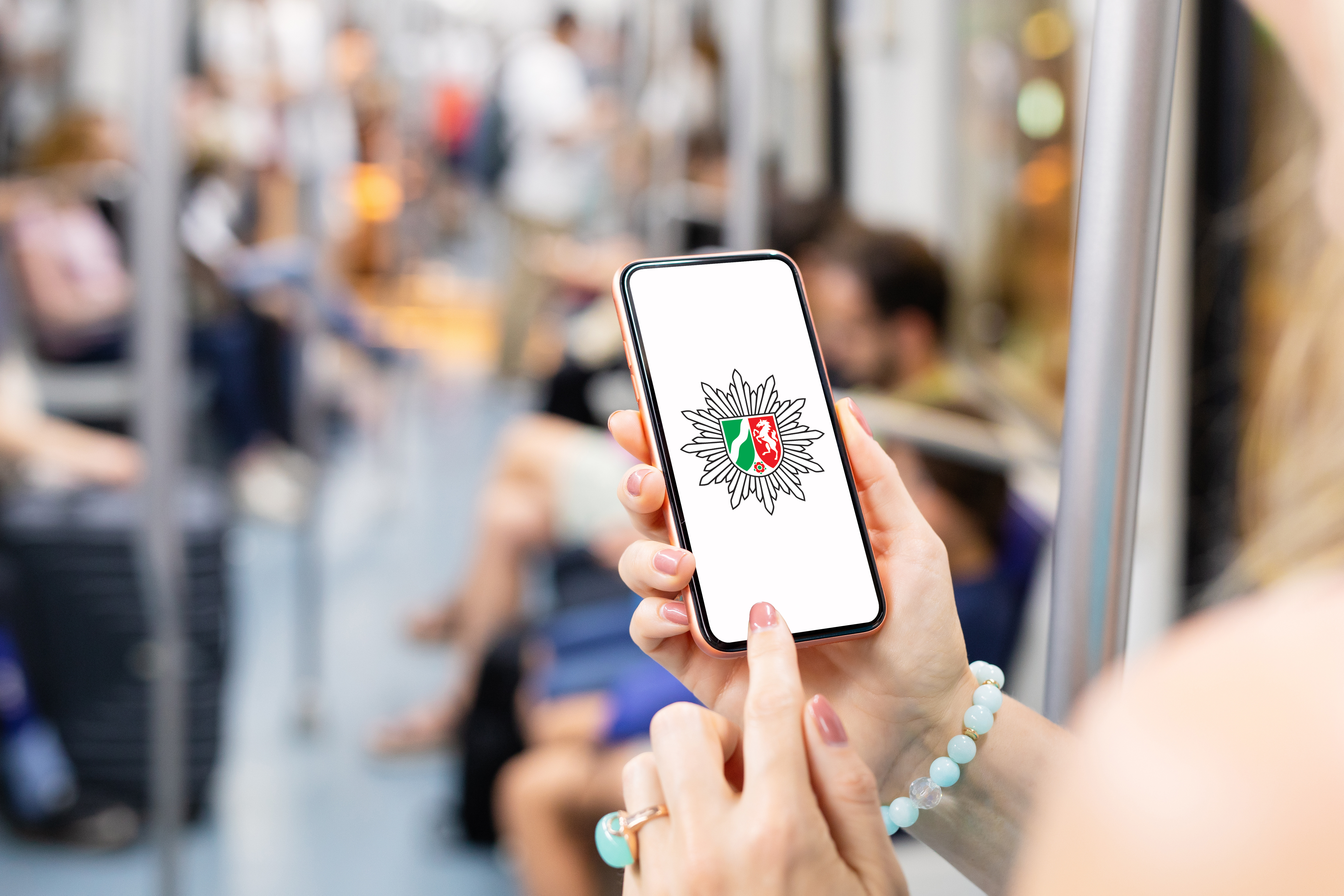 Eine Frau steht in einer U-Bahn und hält ein Handy. Darauf ist das Logo der Polizei NRW zu sehen.