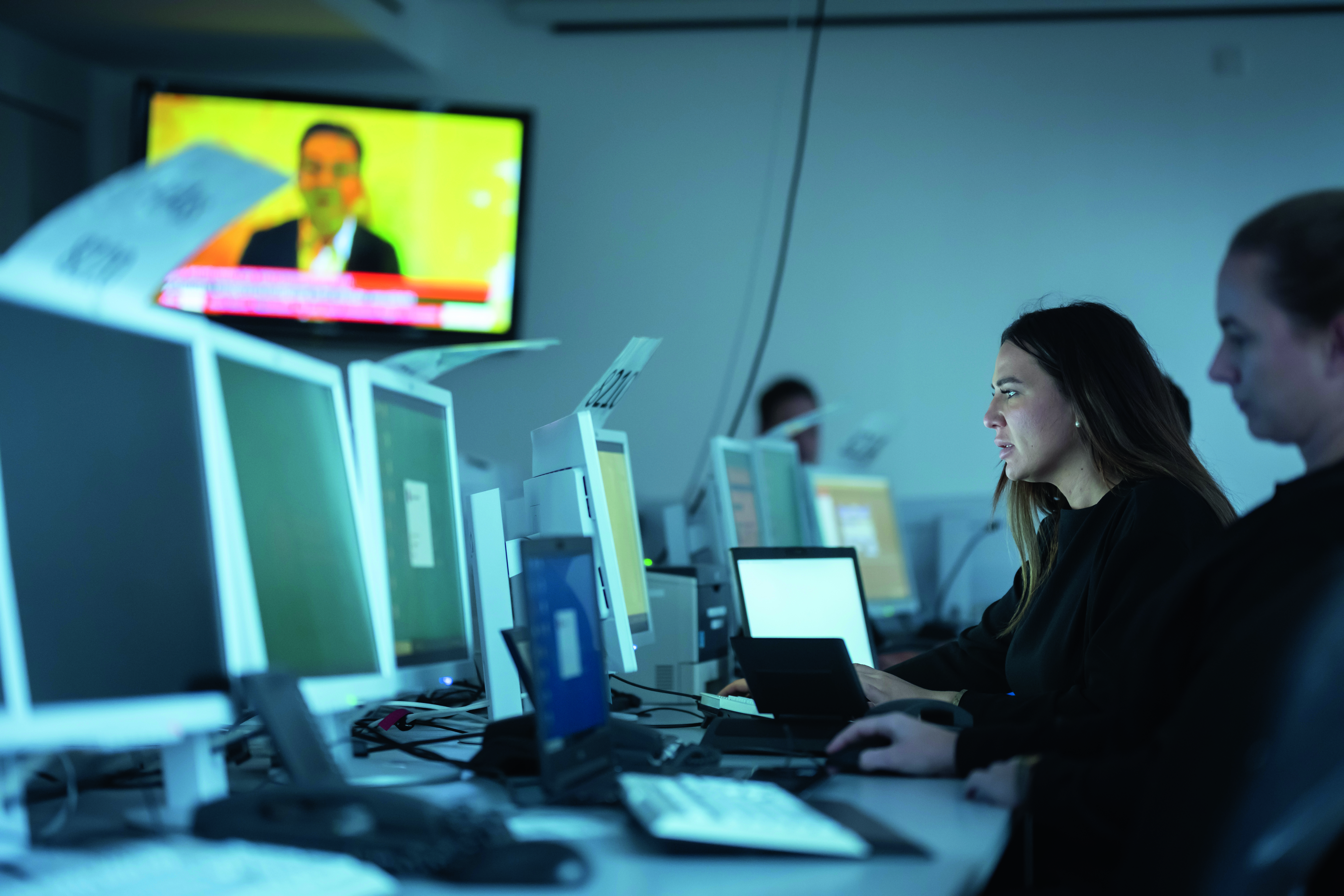 Rechts im Bild sind zwei Frauen zu sehen, die an Bildschirmen arbeiten. Im Hintergrund ist eine weitere Person. Auf dem Arbeitstisch sind viele weitere Bildschirme zu sehen. Oben links ist ein Fernseher zu sehen, auf dem scheinbar Nachrichten laufen.