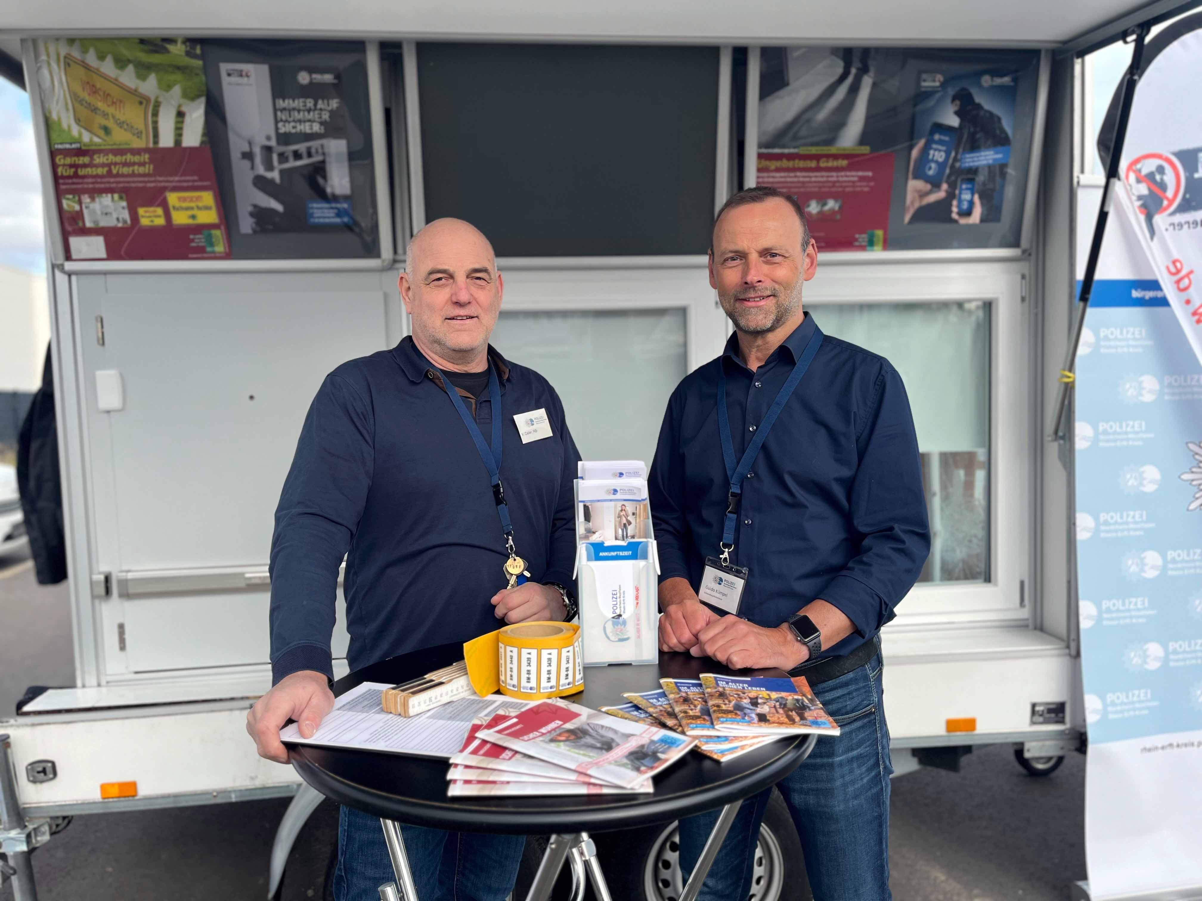 Berater der Kriminalprävention stehen am Marktstand und lächeln in die Kamera. Auf einem Stehtisch vor Ihnen liegt Infomaterial.