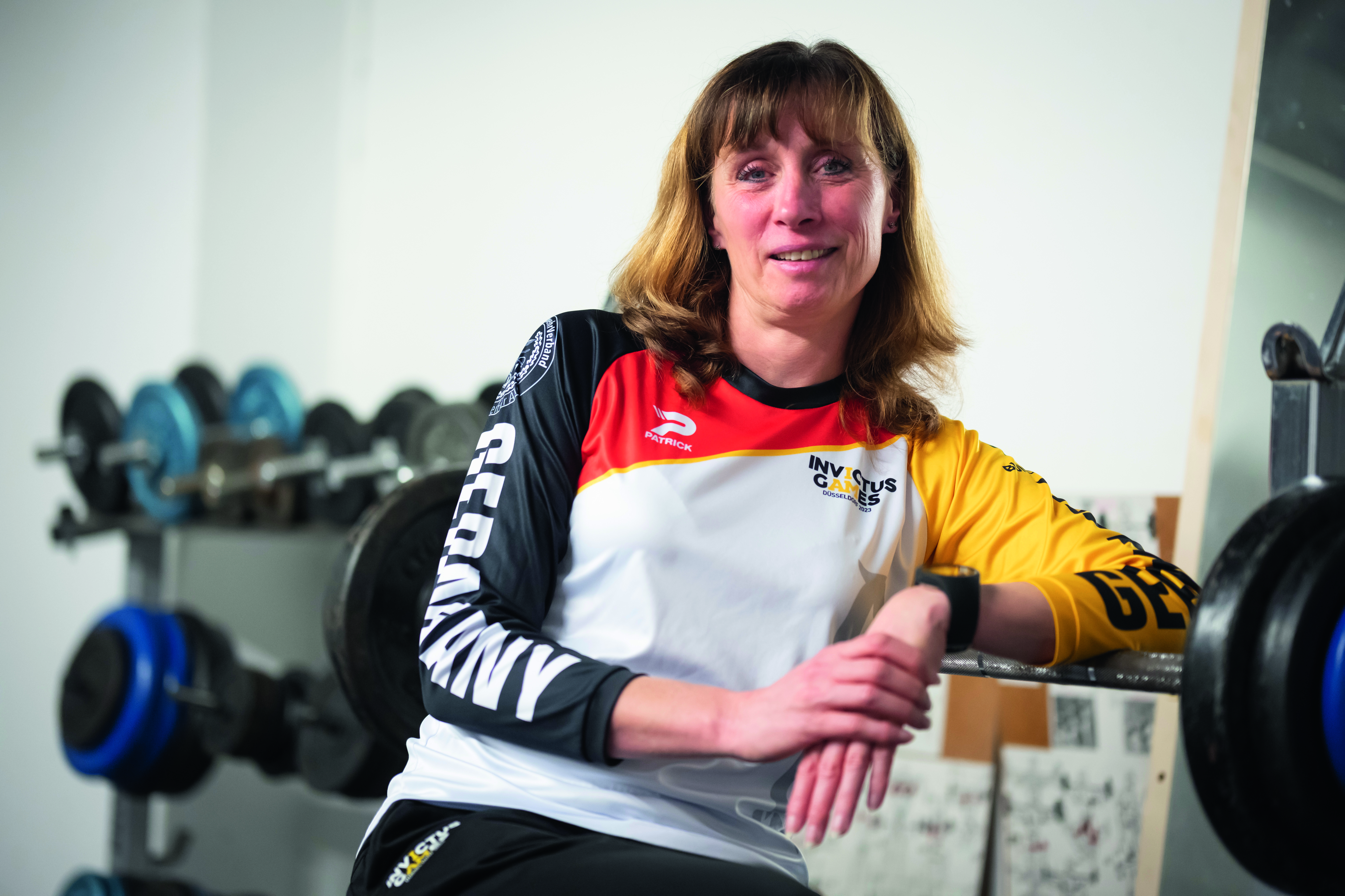 Policewoman Sandra Müller is sitting in the foreground. She is in a training room and is wearing an Invictus Games jersey. Weights on brackets can be seen in the background.