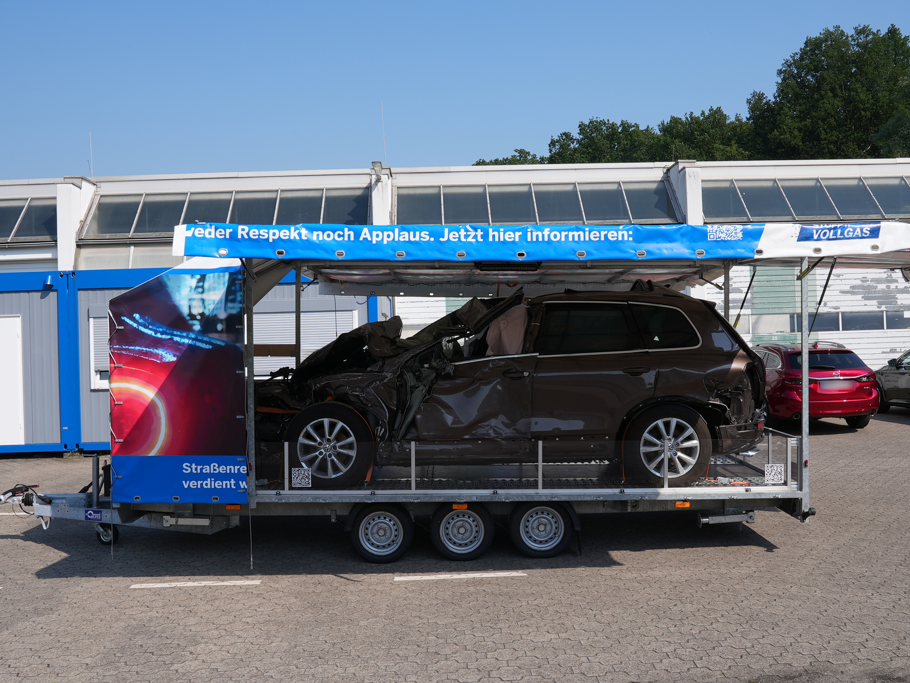 Kampagnenanhänger in Seitenansicht. Die Seitenwände sind hochgeklappt, zu sehen ist ein Fahrzeug mit Totalschaden. Am Dach des Anhängers ist der Schriftzug "Wer Respekt noch Applaus. Jetzt hier informieren" zu sehen