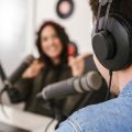 A man and a woman are recording a podcast. The man is seen from behind in the foreground. The woman is seen from the front and is laughing.