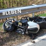 An einer Leitplanke liegt am Straßenrand ein verunfalltes Motorrad auf der linken Fahrzeugseite. Hinter der Leitplanke ist eine steile Böschung mit angrenzendem Laubwald. Auf der Leitplanke steht #LEBEN. Unten rechts steht Symbolbild.