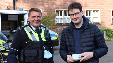 Two people, one in police uniform, are standing next to each other. The person on the right is holding a coffee cup.