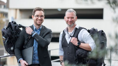 Im Vordergrund sind Kriminaloberkommissar David Berners und Michael Grill zu sehen. Beide tragen schwarze Rucksäcke mit Equipment über einer Schulter. David Berners hält einen Laptop unterm Arm.  Im Hintergrund ist ein helles Parkhaus zu sehen.